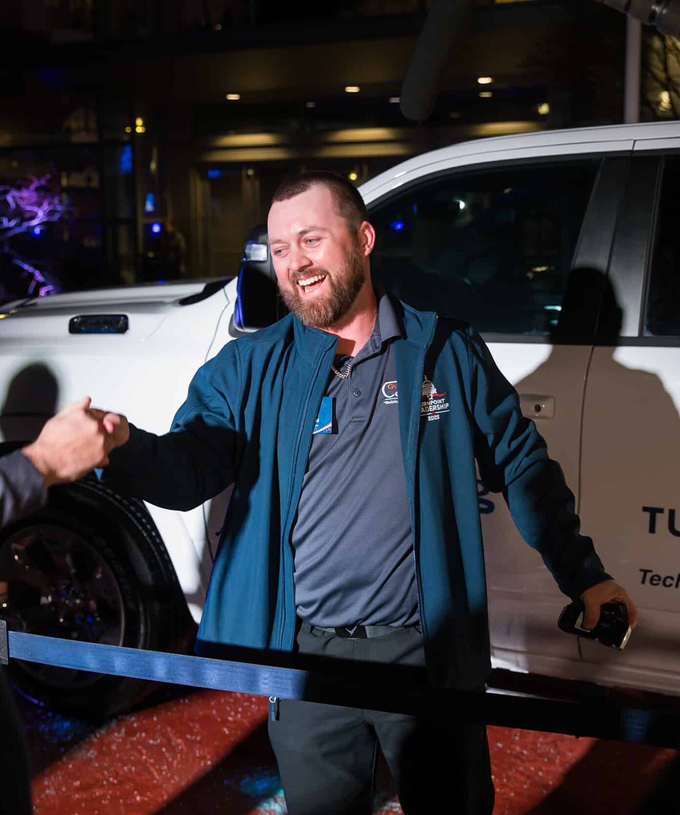 A man wearing a blue jacket shakes hands with someone in front of a white truck labeled TurnPoint Services Technician of the Year at a leadership conference.