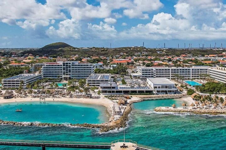 Aerial view of one of the premier all-inclusive resorts in Mexico and the Caribbean, featuring white buildings, pools, palm trees, and a sandy beach surrounded by turquoise water and lush greenery under a partly cloudy sky.