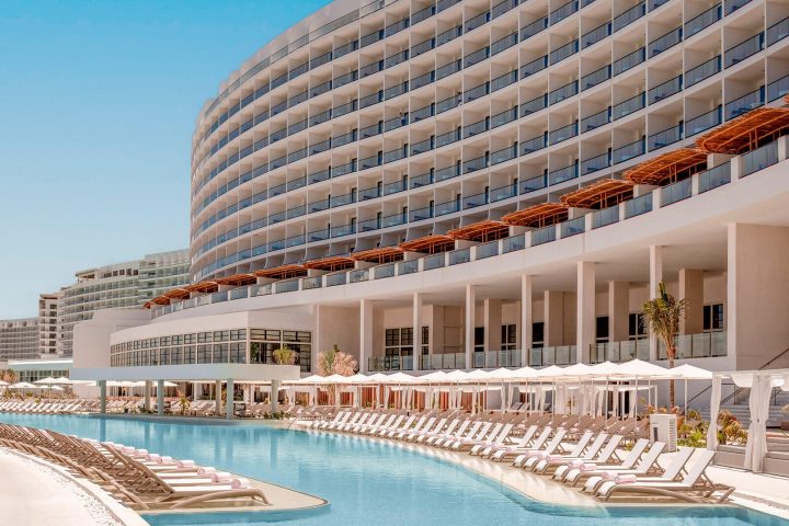 Curved modern hotel building with balconies overlooks a long outdoor swimming pool lined with sun loungers and umbrellas—a scene reminiscent of all-inclusive resorts in Mexico and the Caribbean under a clear blue sky.