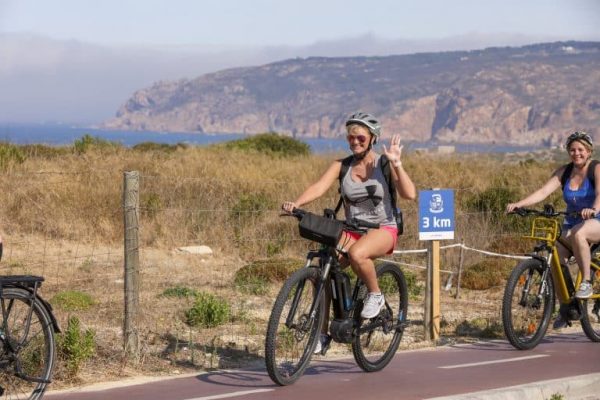 Two women wearing helmets ride bicycles on a paved path near a fence, with dry grass and hills in the background. One woman waves at the camera. A sign reads 3 km—perfect scenery for a Portugal incentive travel adventure.