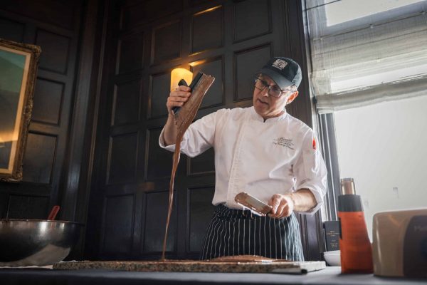 Chef Joel Lahon_DFMotion Photography (1) A chef in a white uniform and hat pours melted chocolate onto a countertop from a scraper in a dimly lit kitchen, capturing the sweet allure that makes Incentive Travel to Québec City unforgettable.