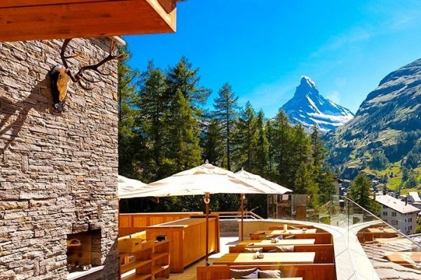 Madre-Nostra-202210-BL Outdoor terrace with wooden seating and umbrellas overlooks an alpine village and trees, with the Matterhorn mountain visible under a clear blue sky—a perfect spot featured in the Switzerland incentive travel guide.