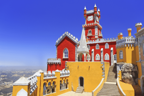 A brightly colored castle with red, yellow, and gray walls, featuring towers and staircases, sits under a clear blue sky—an iconic sight perfect for Portugal incentive travel with its inspiring distant landscape.