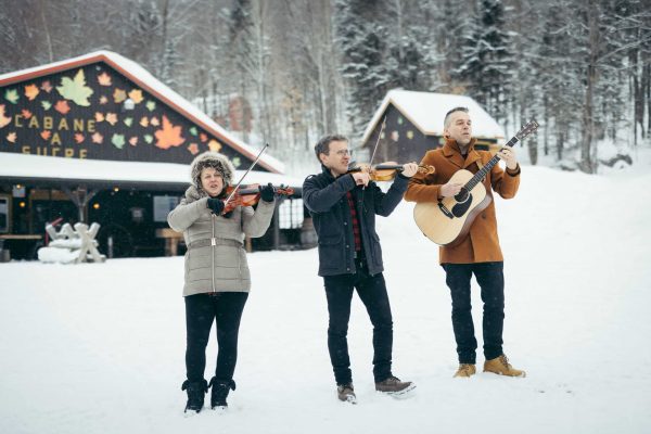 Sugar Shack_DFMotion Photography Three people play music outside in the snow; two play violins and one plays an acoustic guitar, creating a magical scene perfect for Incentive Travel to Québec City, with a cabin and snowy trees in the background.
