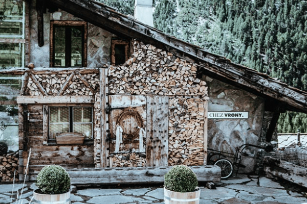 chez vrony A rustic wooden cabin with stacked firewood, small windows, and two potted shrubs in front; set against a forested mountain backdrop—straight from a Switzerland incentive travel guide.