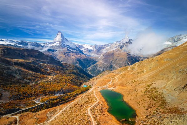 Zermatt, Switzerland with the Matterhorn and Stellisee Lake A mountain landscape with a green alpine lake, winding trails, autumnal trees, and snowy peaks like the Matterhorn beneath a partly cloudy blue sky—perfect inspiration for any Switzerland incentive travel guide.